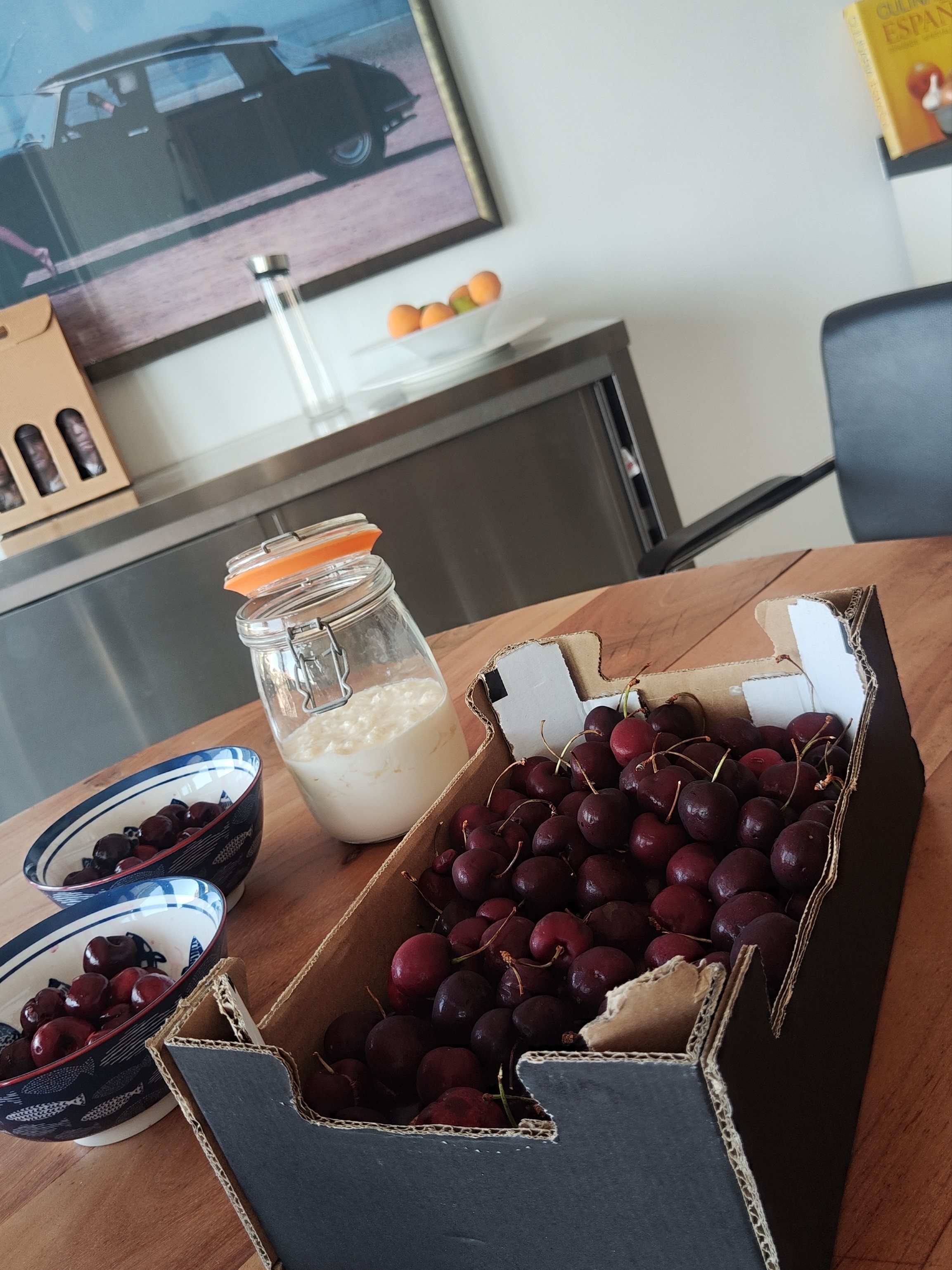 Round wooden kitchen table with a crate of two kilos of cherries, two bowls of pitted cherries, and a jar of homemade fermented kefir. In the background, a poster of a classic Citroën DS above a stainless steel cabinet with a fruit bowl full of citrus and the cookbook Culinaria España.