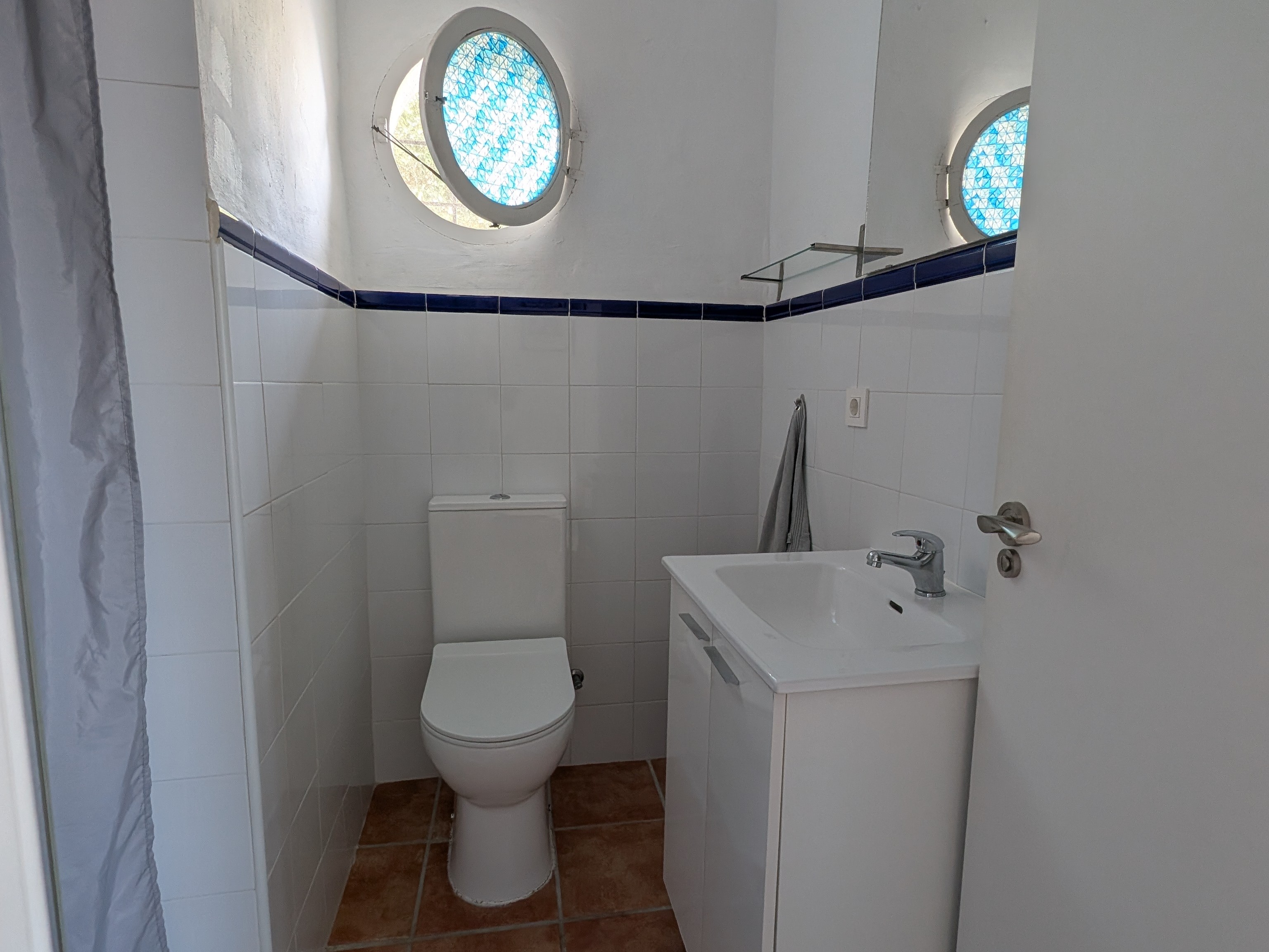 De kleine maar praktische badkamer van Villa Merise, betegeld in witte en blauwe Spaanse retrostijl. Op deze foto zie je het toilet, de wastafel met spiegel en het kleine ronde schippersraam dat zorgt voor natuurlijk licht
