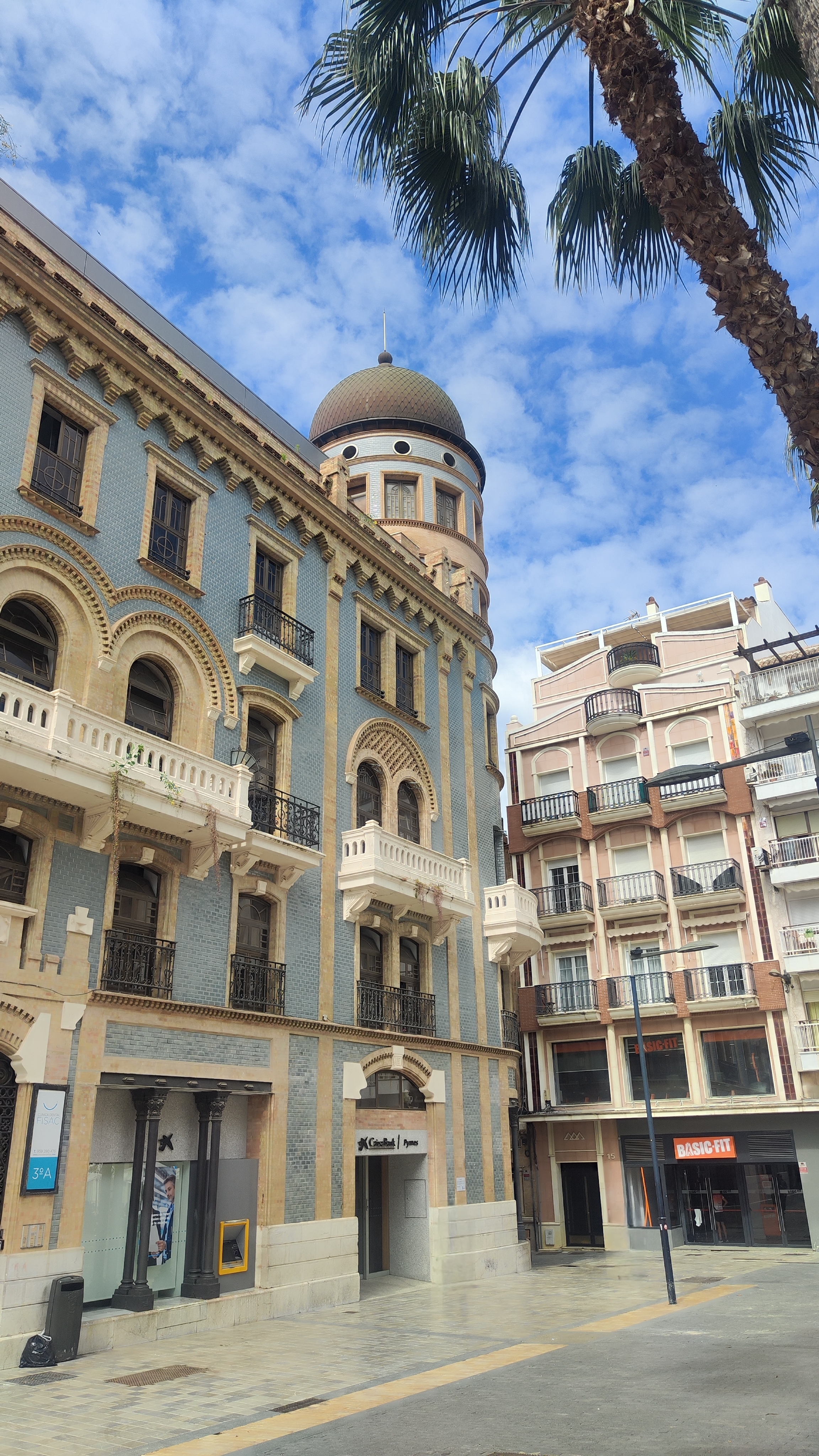 Het blauwe Belle Époque-gebouw met een ronde toren aan een plein met marmeren bestrating in Huelva. Op de foto is alleen de hoek met de toren zichtbaar, samen met een palmboom en een blauwe lucht met lichte wolkjes.