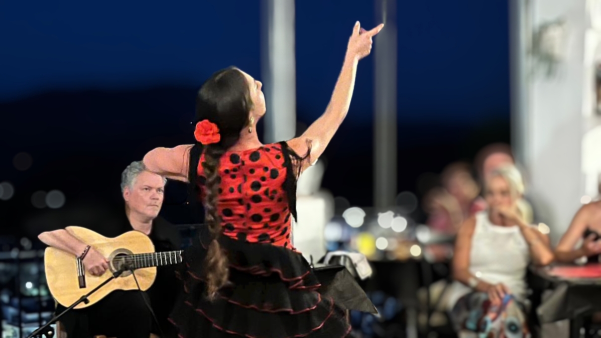 Victoria Macías Costela, flamencodanseres in een rood-zwarte jurk met zwarte polka dots, dansend in Restaurante La Traviesa. Ze kijkt en wijst omhoog, terwijl op de achtergrond de gitarist speelt.