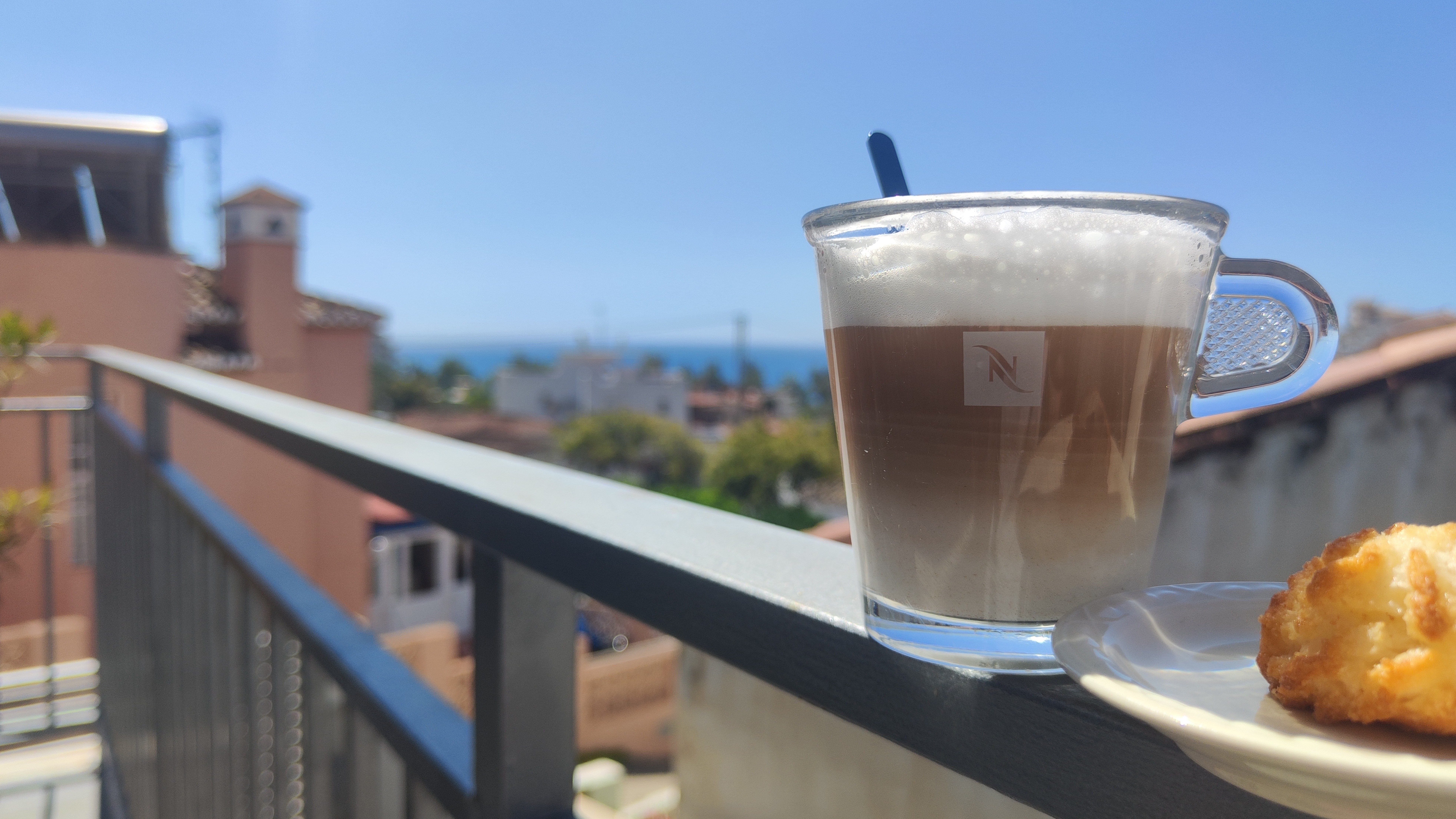 Nespresso-glas met café manchada en kokosmakroon op balkonrand met uitzicht op baai van Pozuelo en park in Almuñécar, Andalusië