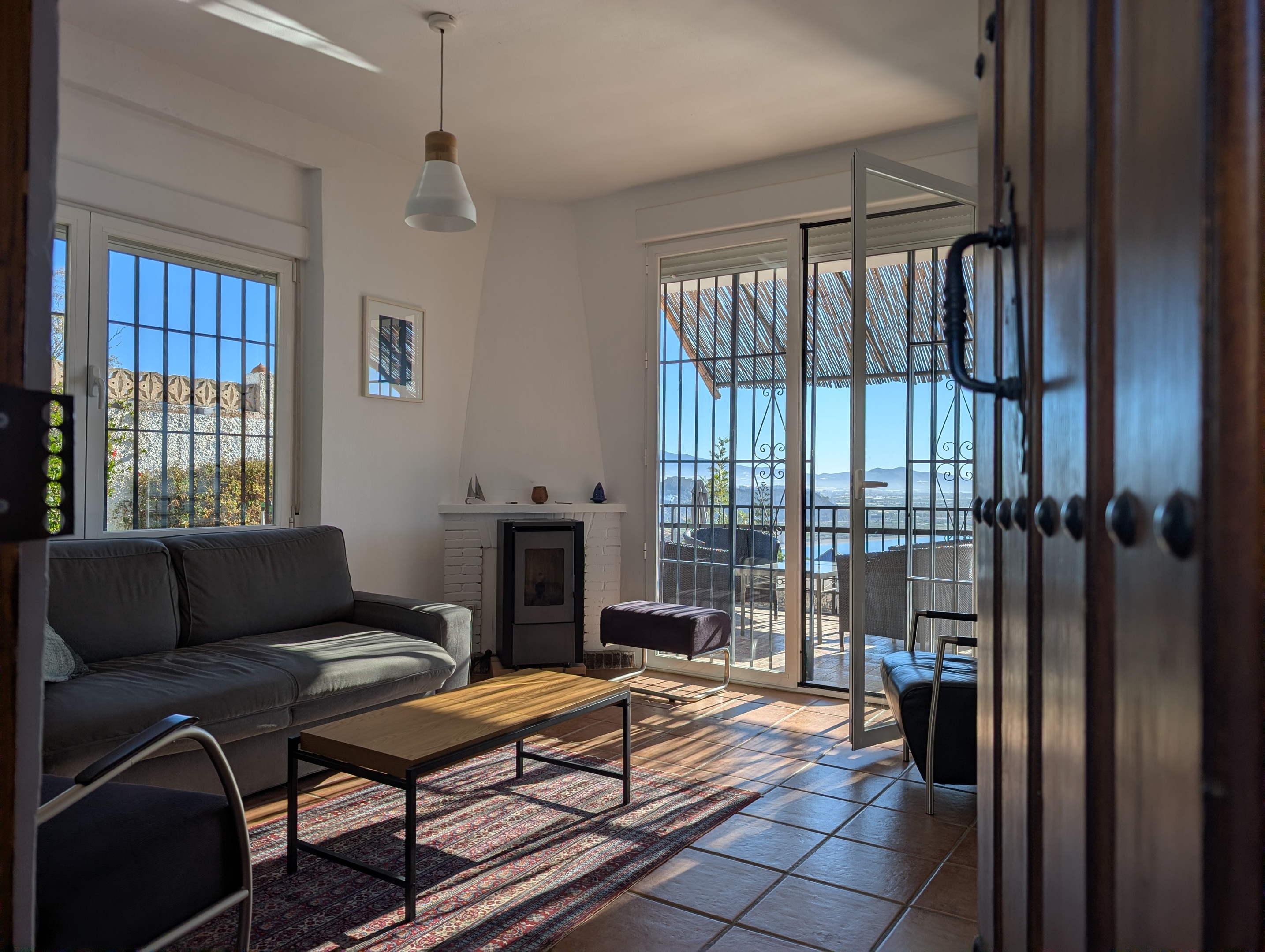 Binnenkomst door de rustieke houten paneeldeur van Villa Merise, waar je direct het panoramische uitzicht op de veranda, zee en bergen kunt zien. De openslaande deuren brengen je naar een prachtig buitenruimte, met de horizon van de Middellandse Zee en het omliggende berglandschap