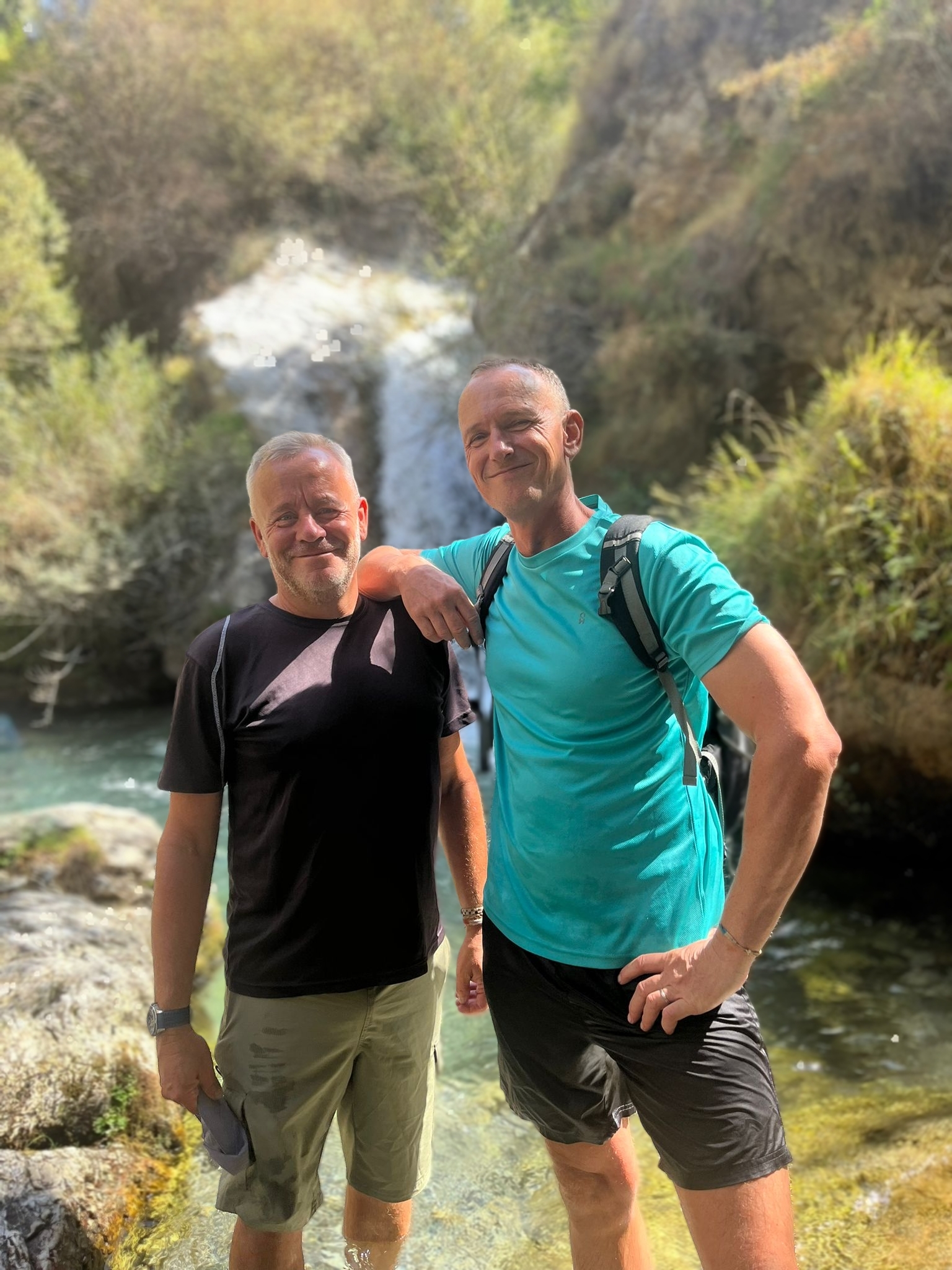 Johan en Reijer in sportieve outfits bij de waterval van de Río Dilar, lachend en vriendelijk in de camera, zichtbaar gelukkig.