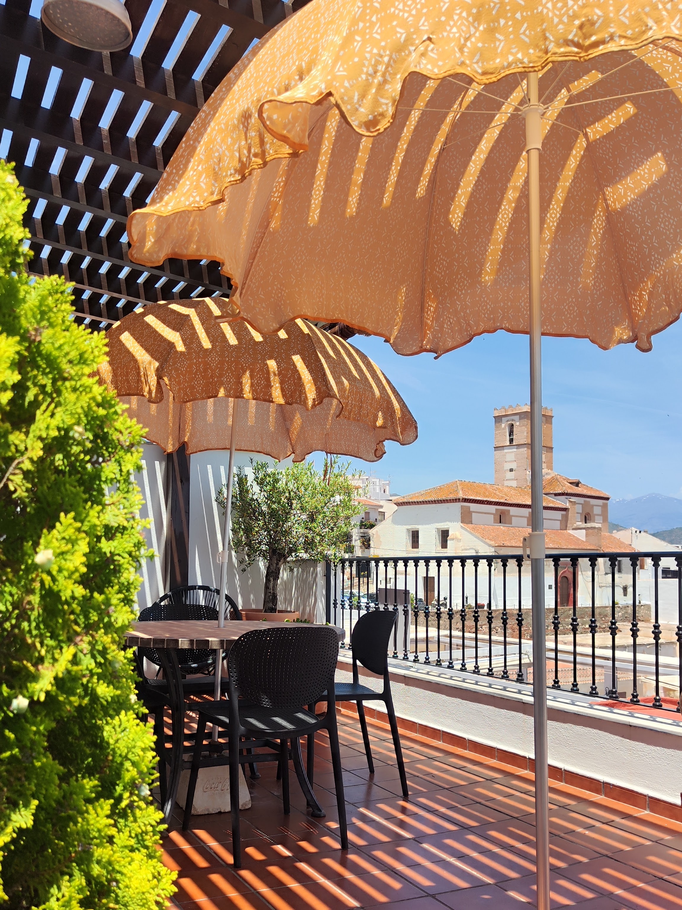 View from the terrace of Ático Lolapaluza under the pergola with two orange Ibiza-style parasols, dining table, olive trees and terracotta pots, overlooking the church and the village of Salobreña, with the Sierra Nevada in the background in summer.
