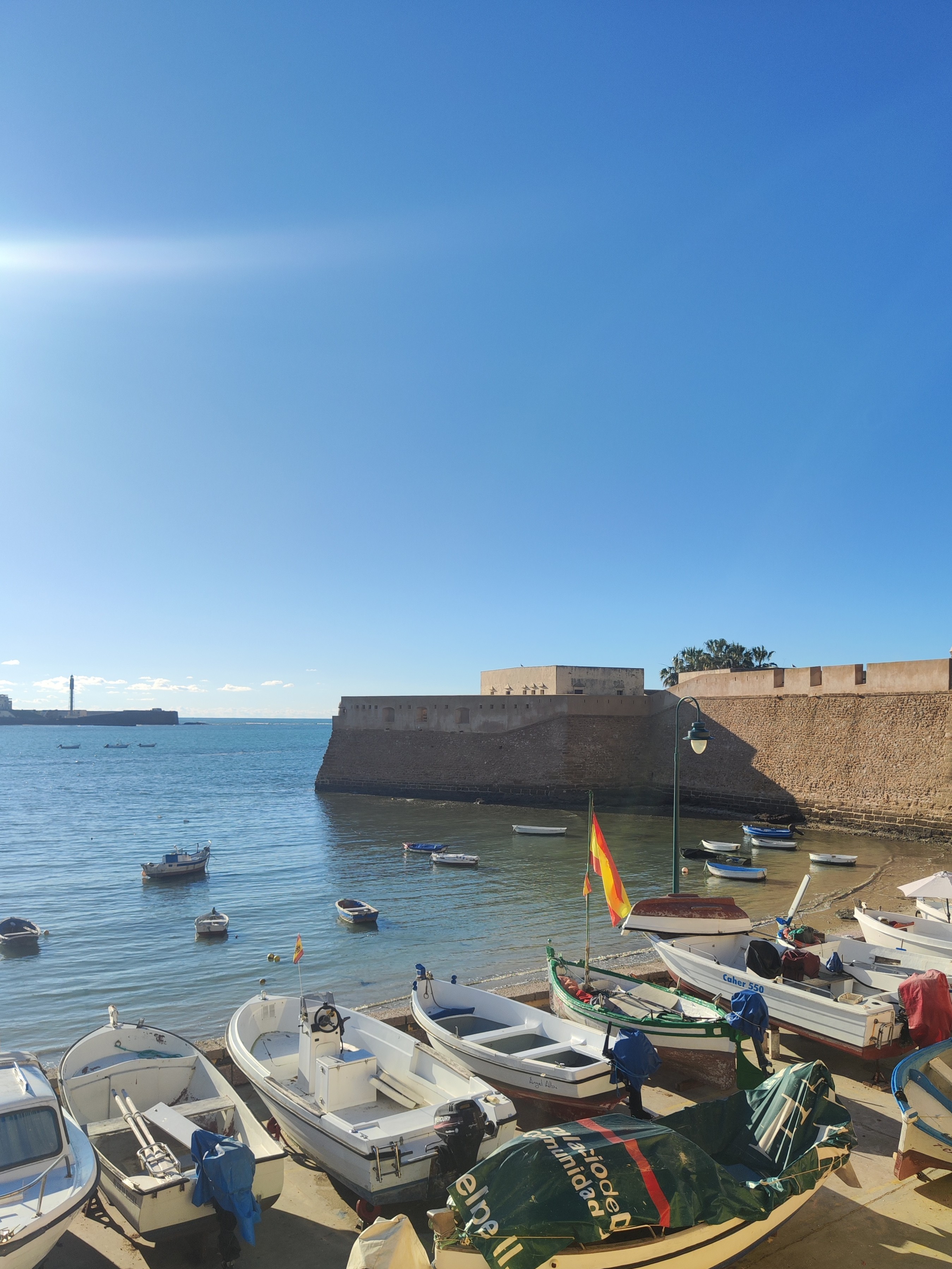 Haven en strand van Cádiz met bootjes, fort en pier bij kalme zee en zilver zonnige lucht