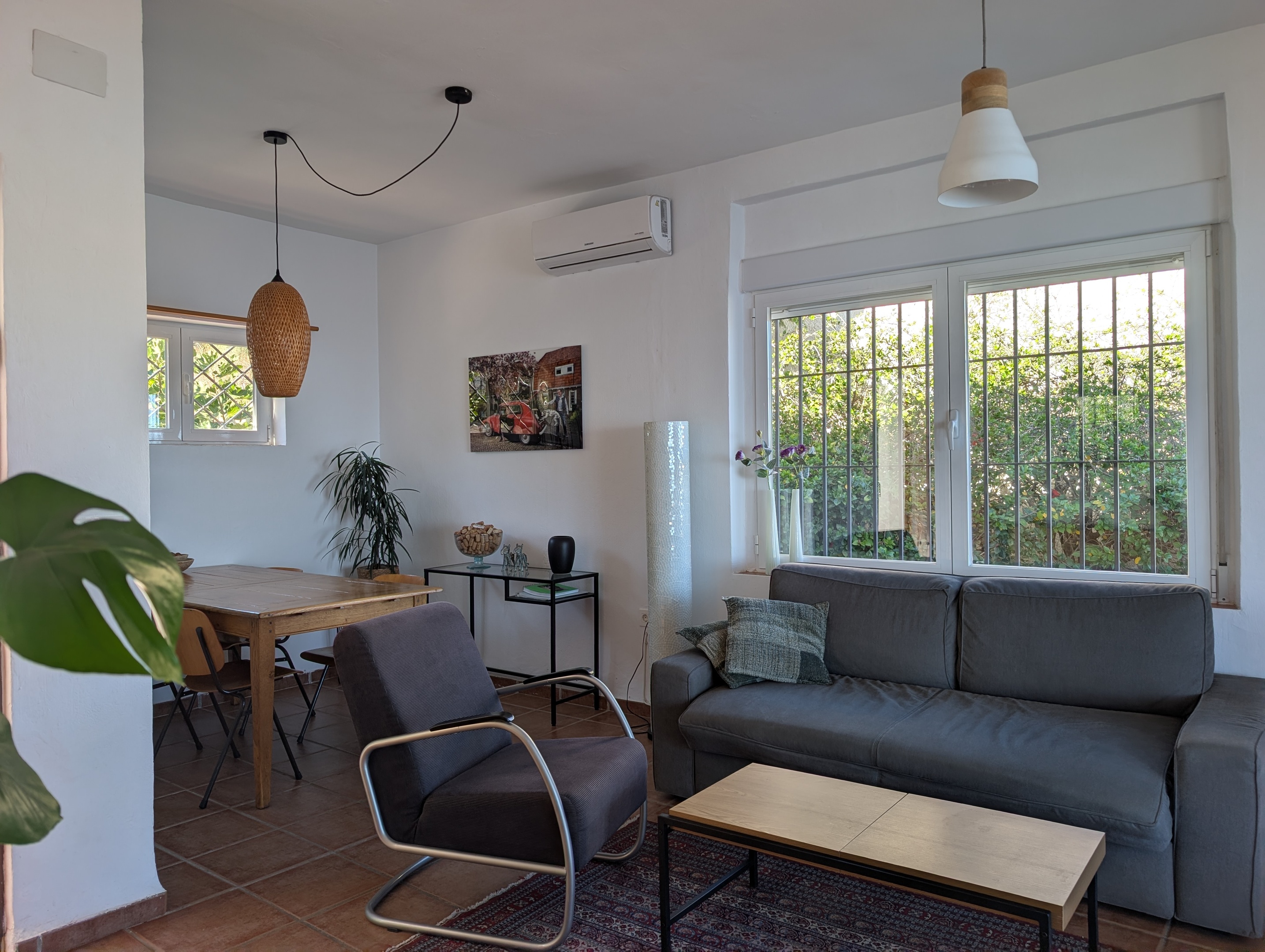 De zithoek van Villa Merise met retro fauteuils, een tweepersoons zitbank die als bed gebruikt kan worden, en een perzich tapijt. De pelletkachel biedt warmte voor de koelere avonden, terwijl de airco zorgt voor zowel verwarming als koeling. Het grote raam biedt uitzicht op een rood bloeiende hibiscus. De villa ligt in de buurt van Salobreña en biedt makkelijk toegang tot de Costa Tropical.