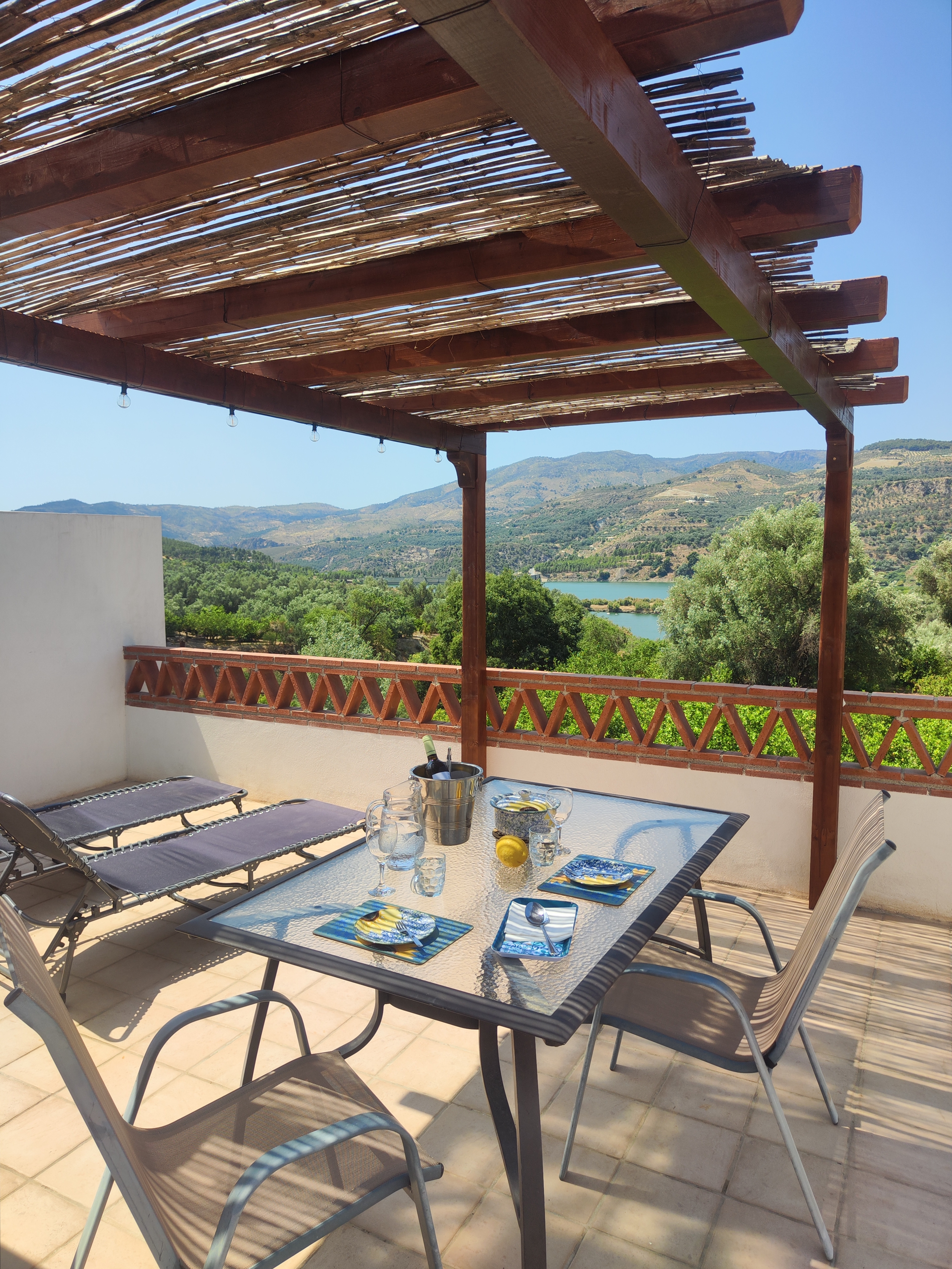 Pergola with a reed roof on the rooftop terrace of Casita Klein Zwitserland, with a set table and two sunbeds. View over the terrace, orange groves, Lake Beznar, and hills with pine trees. Terracotta tile terrace railing in a house-of-cards pattern, typical of the Alpujarras.