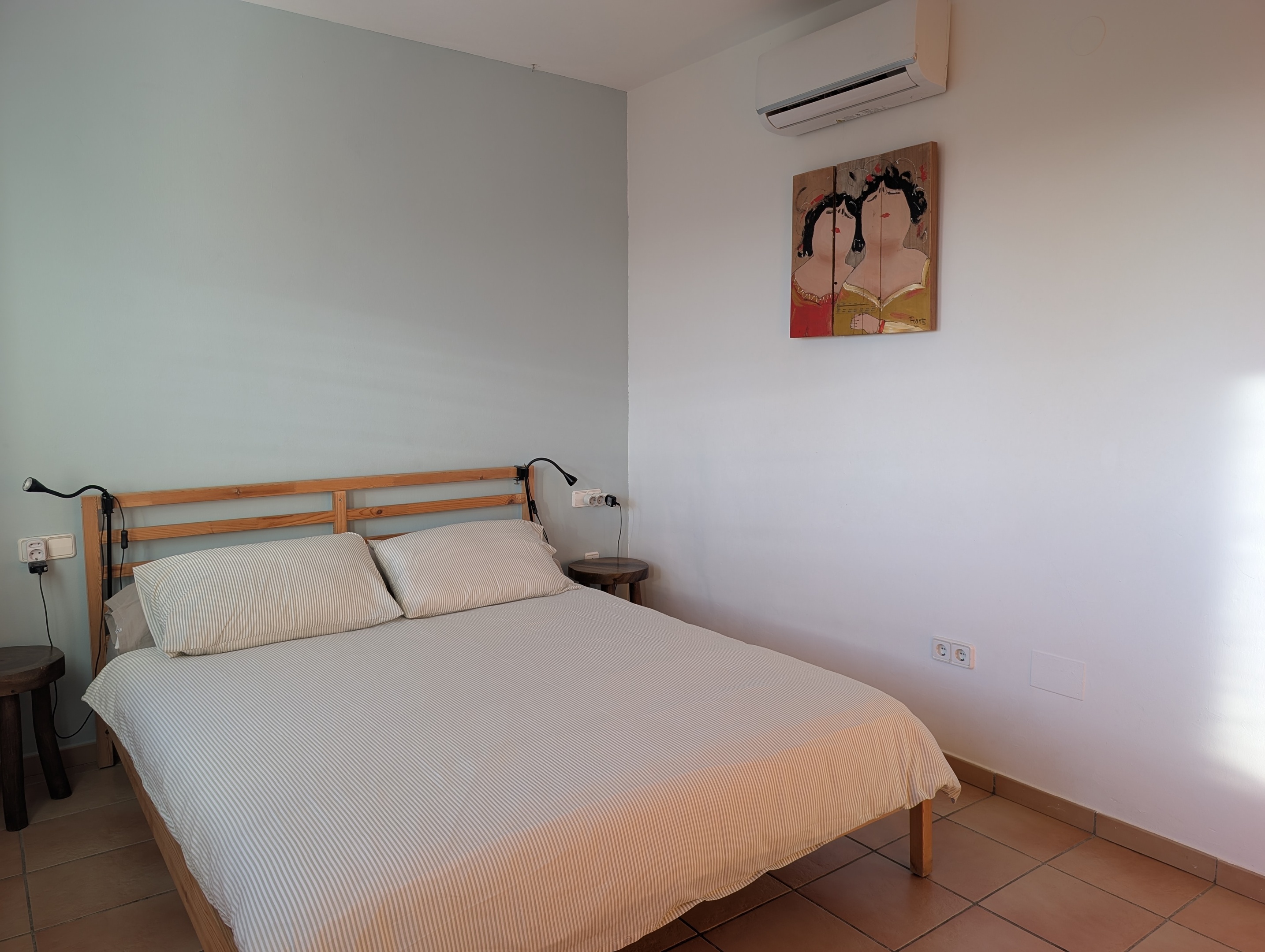 Comfortabel tweepersoonsbed in de masterbedroom van Casa Larimar, met kunst van 'Dikke Dames' op wrakhout, geïnspireerd door de kleuren Geel en Rood van Spanje. Boven het bed bevindt zich een airco voor koelen en verwarmen.