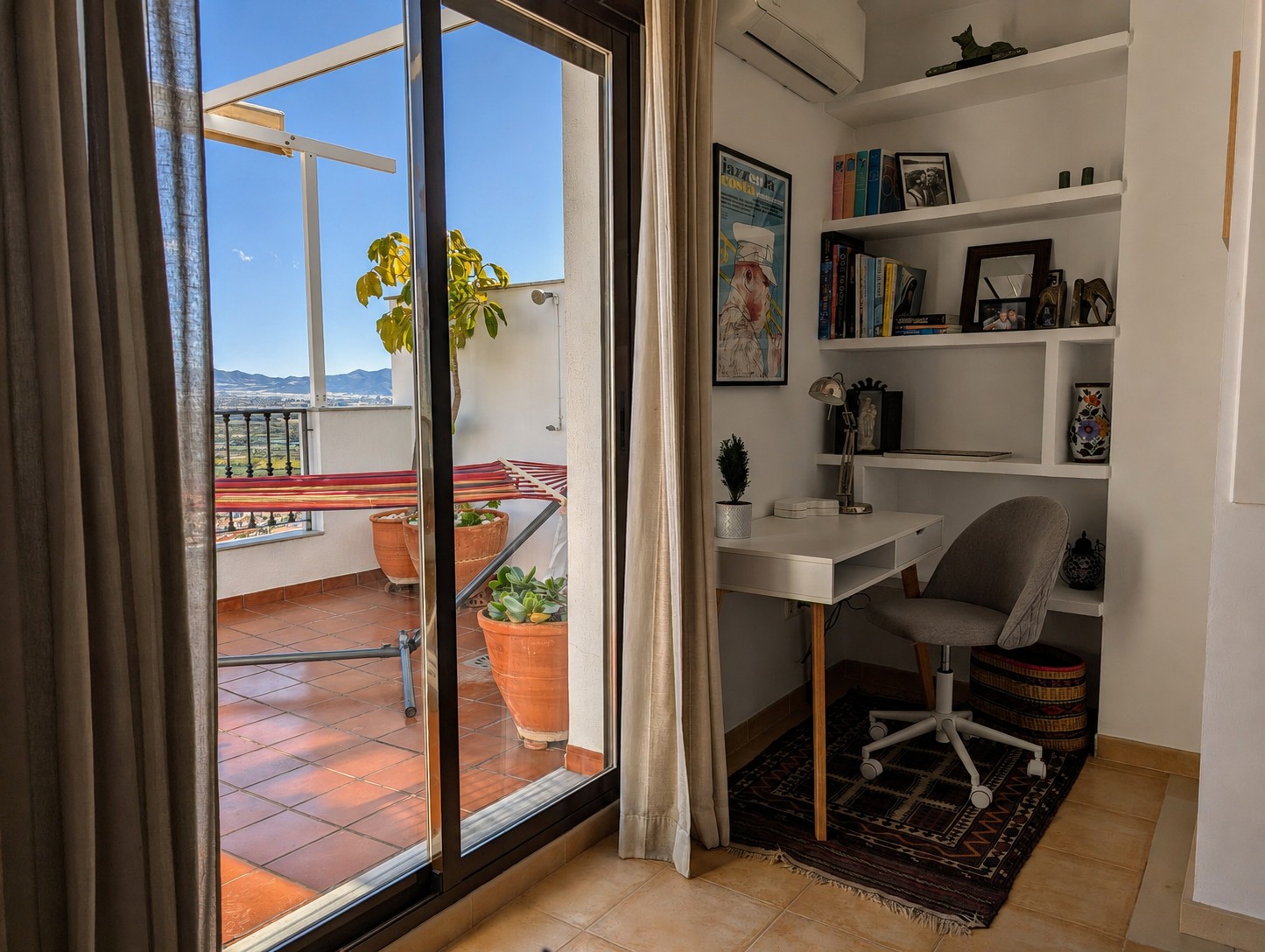 Werkplek in Ático Lolapaluza met schuifpui naar het dakterras, potplanten, hangmat en kleurrijke reis- en architectuurboeken. Boven de werkplek de airco, met Spaanse en Afrikaanse reisdetails en muurprenten uit Almuñécar en Salobreña