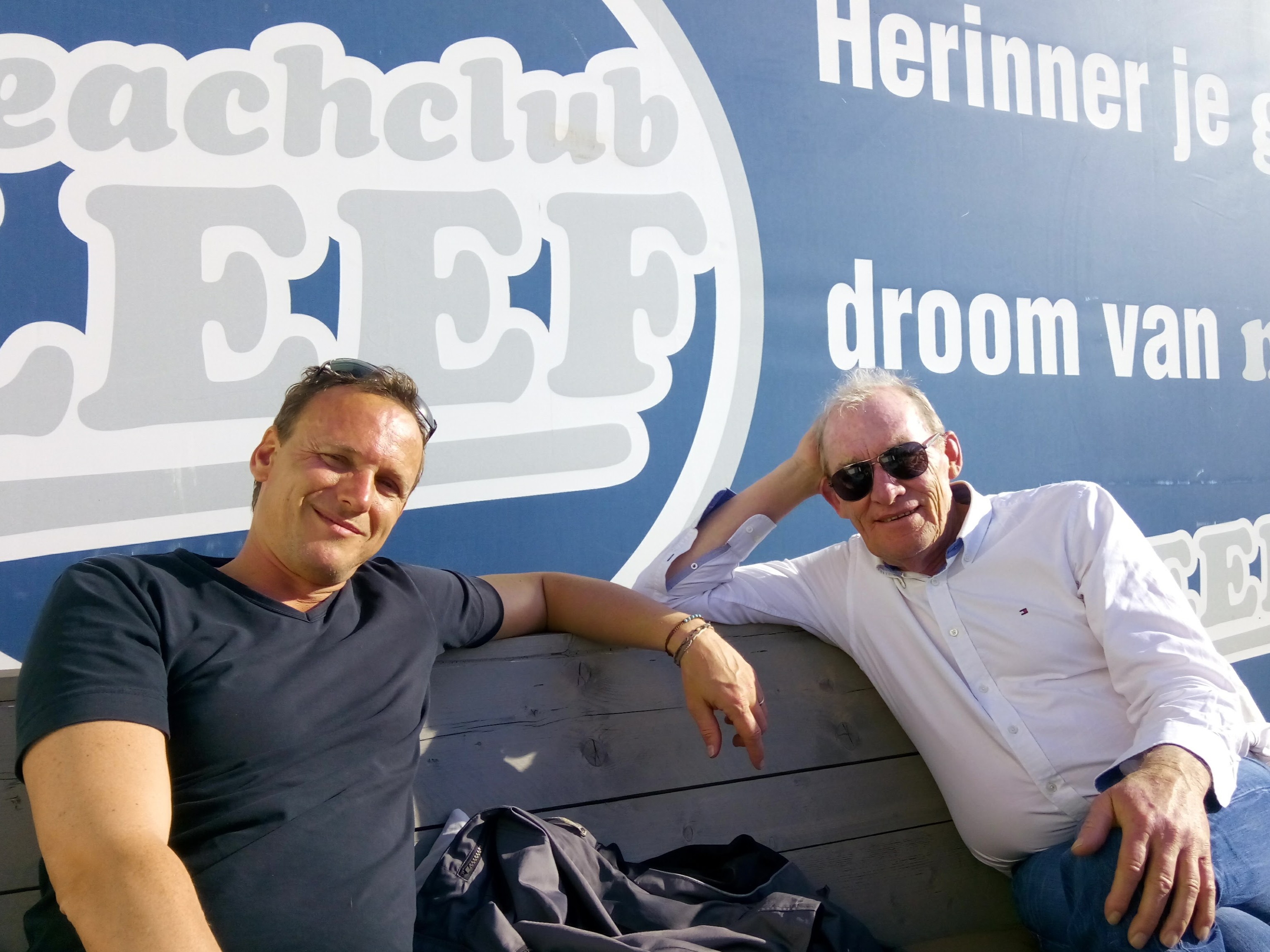 Reijer en Simon in 2017 bij Beachclub LEEF in Kijkduin, poserend voor een blauwe achtergrondwand met het logo en de tekst 'Leef vandaag, Herinner je gister, droom van morgen'.