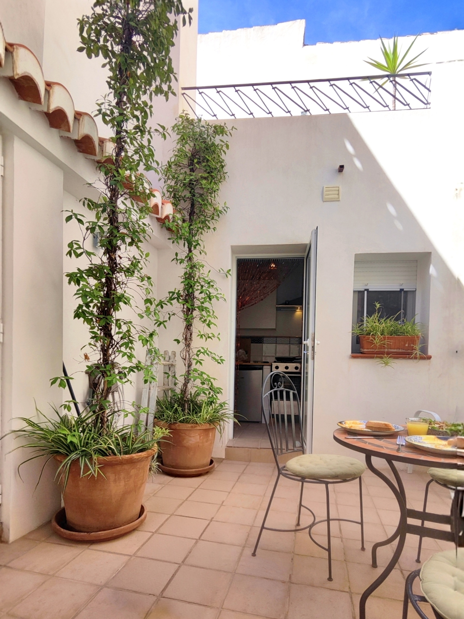 De deur tussen de keuken en de patio van Casita Klein Zwitserland, gezien vanuit de patio. Jasmijn en een graslelie sieren de vensterbank, terwijl het kralengordijn een speels accent toevoegt. Op het tussen-dakterras is authentiek ijzerwerk te zien.