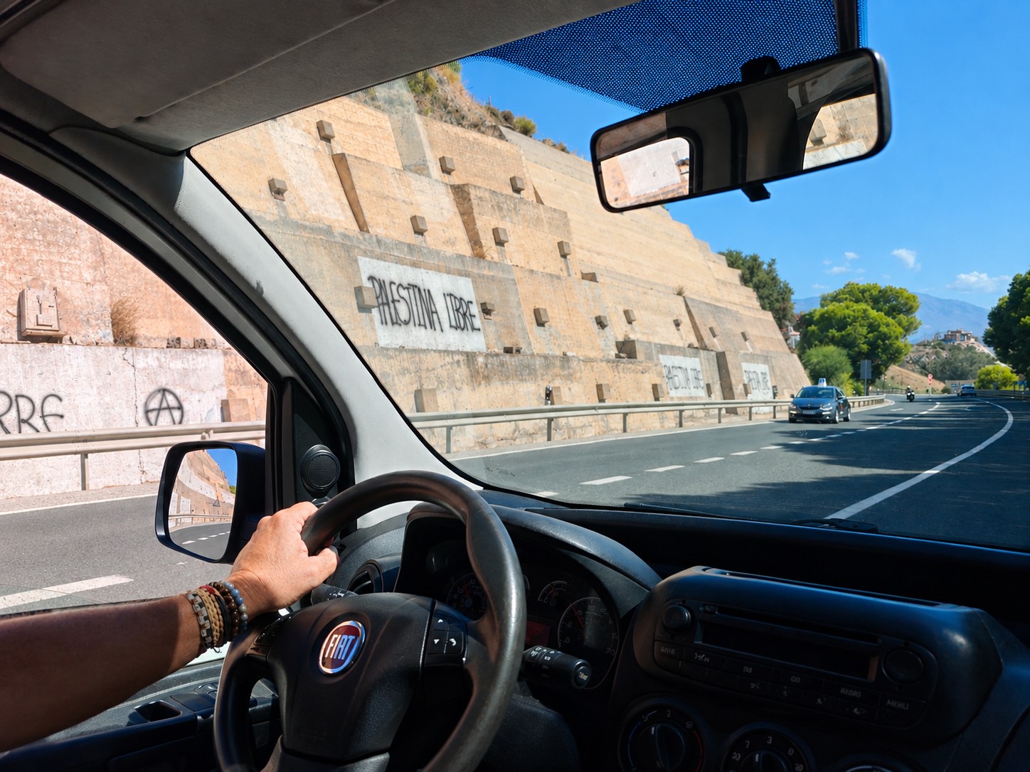 Uitzicht vanuit een rijdende Fiat op de Spaanse kustweg N-340 richting Salobreña, met aan de bergzijde een hoge steunmuur van betonblokken voorzien van graffiti Palestina Libre en in de verte de bergen onder een blauwe lucht.