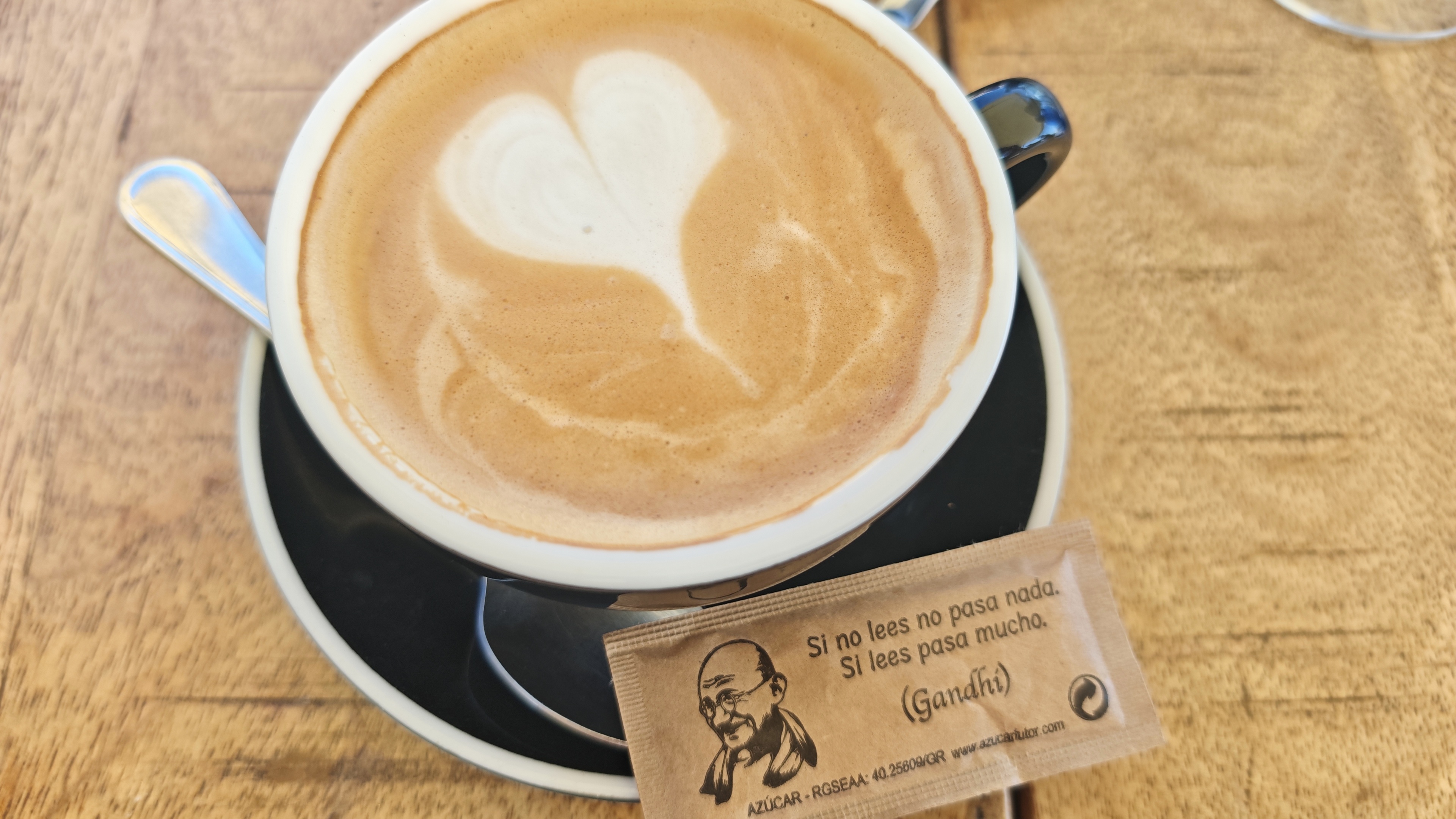 Een vers gemaakte cappuccino in een zwarte kop met latte art in de vorm van een hartje, geserveerd op een houten tafel met een bruin suikerzakje met een citaat van Gandhi in het Spaans.