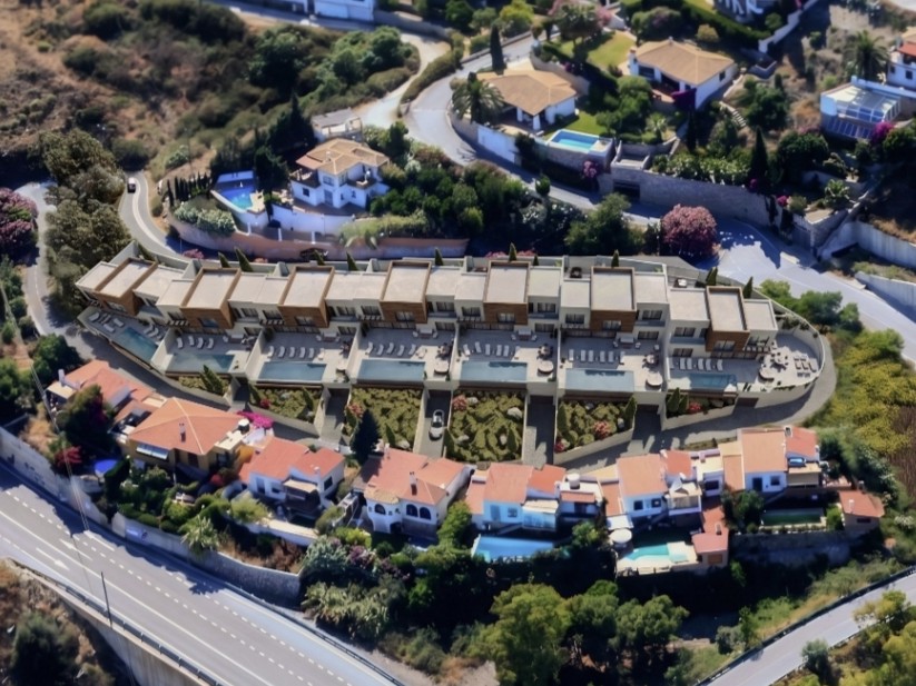 Luchtfoto van de urbanisatie Costa Aguilera Alta in Salobreña aan de kustweg N-340. Centraal op de heuvel staat het moderne geschakelde nieuwbouwcomplex Horizon Costa Aguilera met witte gevels en privé-overloopzwembaden. Direct daaronder ligt de oorspronkelijke rij mediterrane villa's aan de bergzijde van de kustweg.