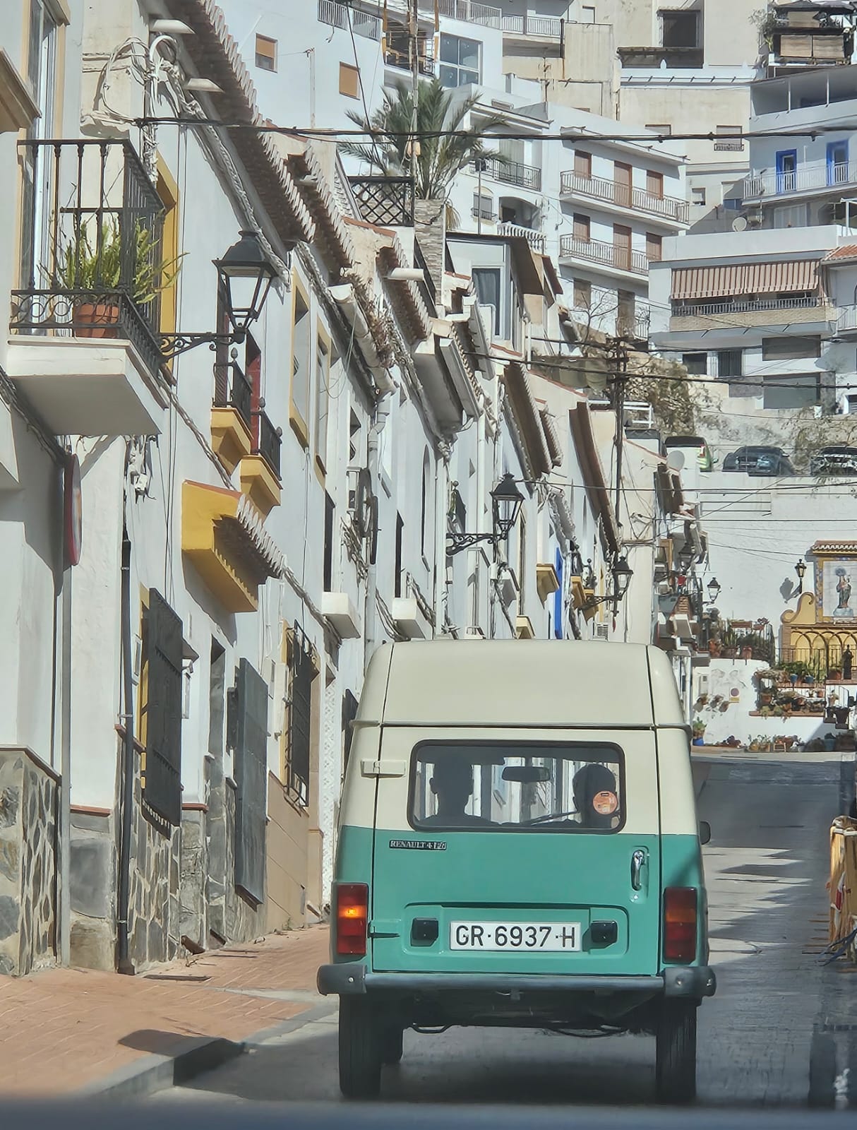 Renault 4 Fourgonnette driving uphill on the steep, cobblestone-paved Calle Rosario in the historic centre of Salobreña. The street is lined with white façades and ends at a Mediterranean-style statue of Mary.